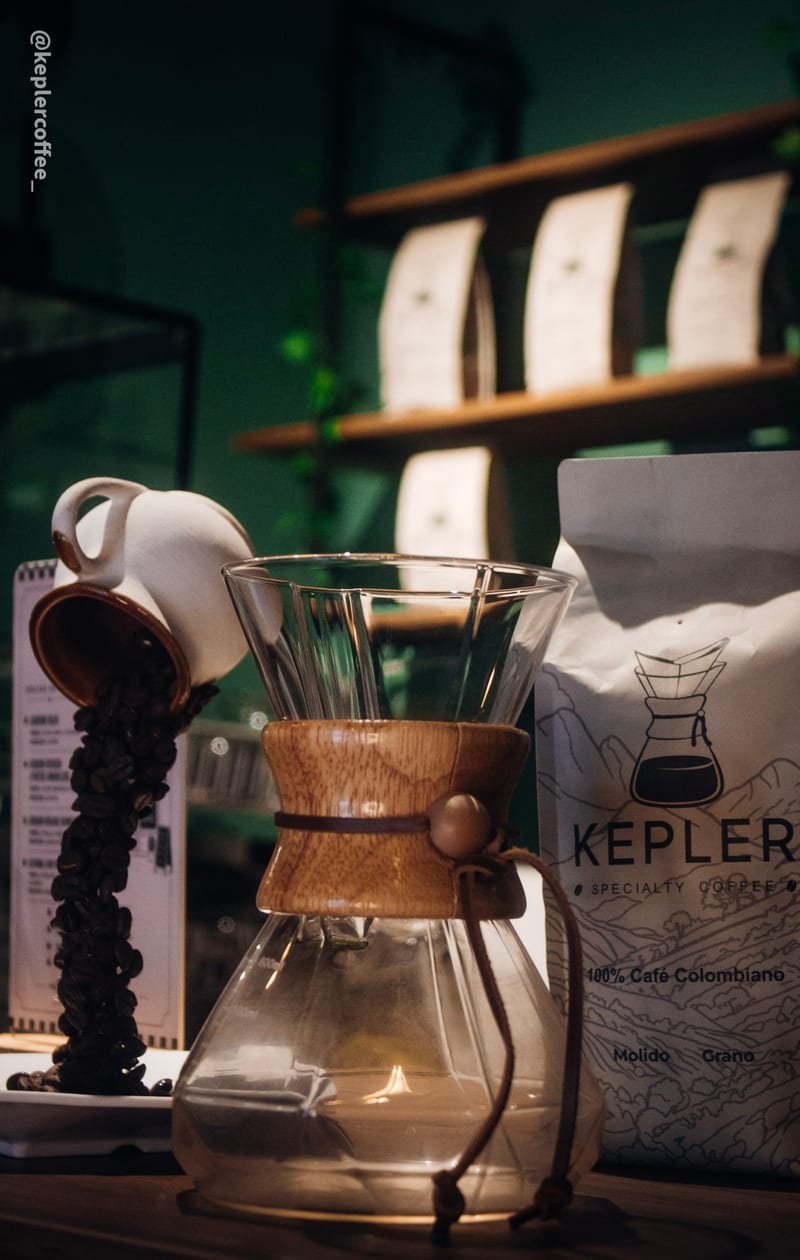 Chemex de vidrio con collar de madera en la barra de Kepler, junto a una bolsa de café de la casa y una taza volcada derramando granos